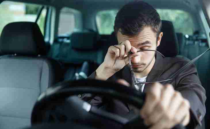 sleepy young man rubs his eyes with his right hand. his left hand is on the steering wheel. he is sitting at his car. road safety concept.